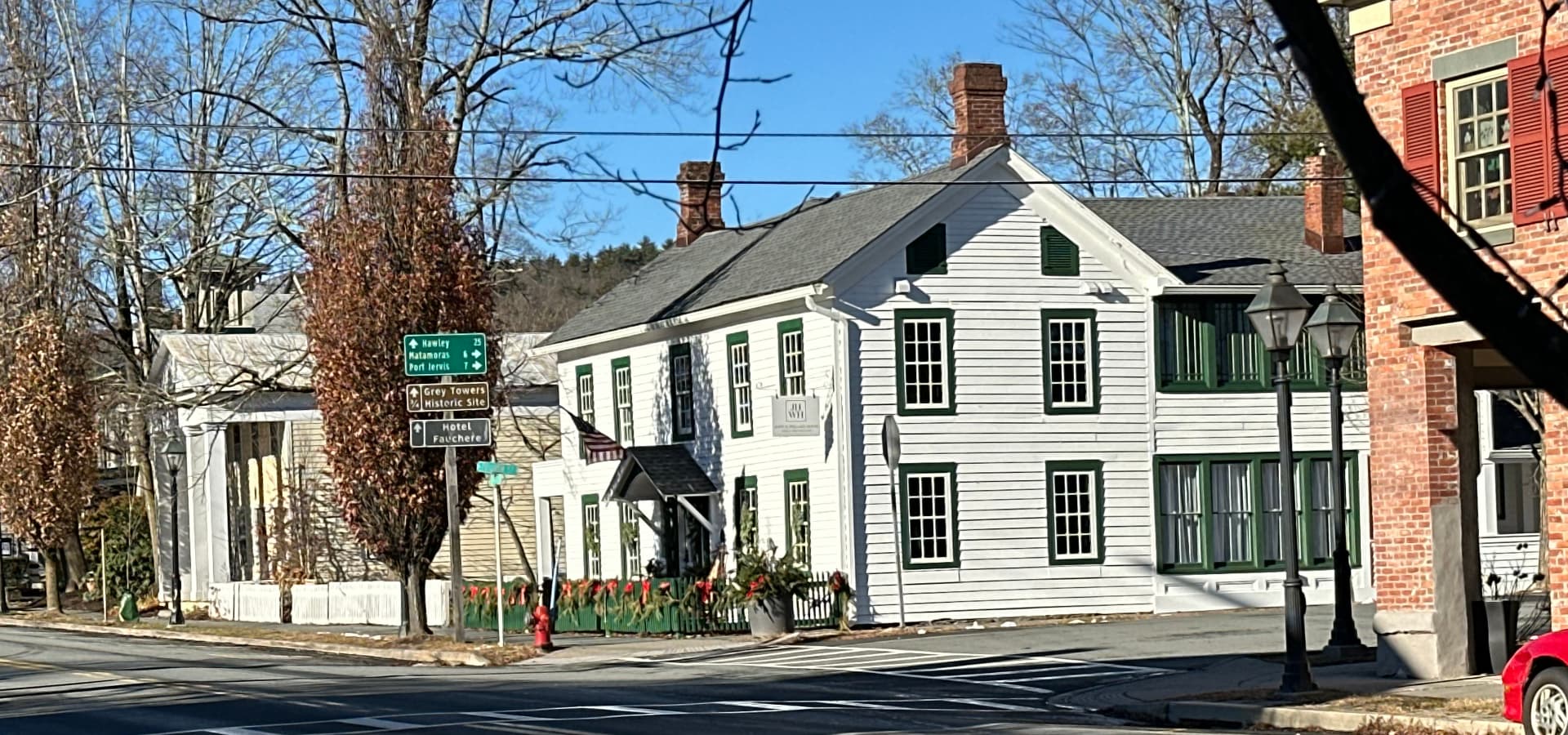 A quaint, white building with green trim is situated at a corner intersection, surrounded by trees and street signs.