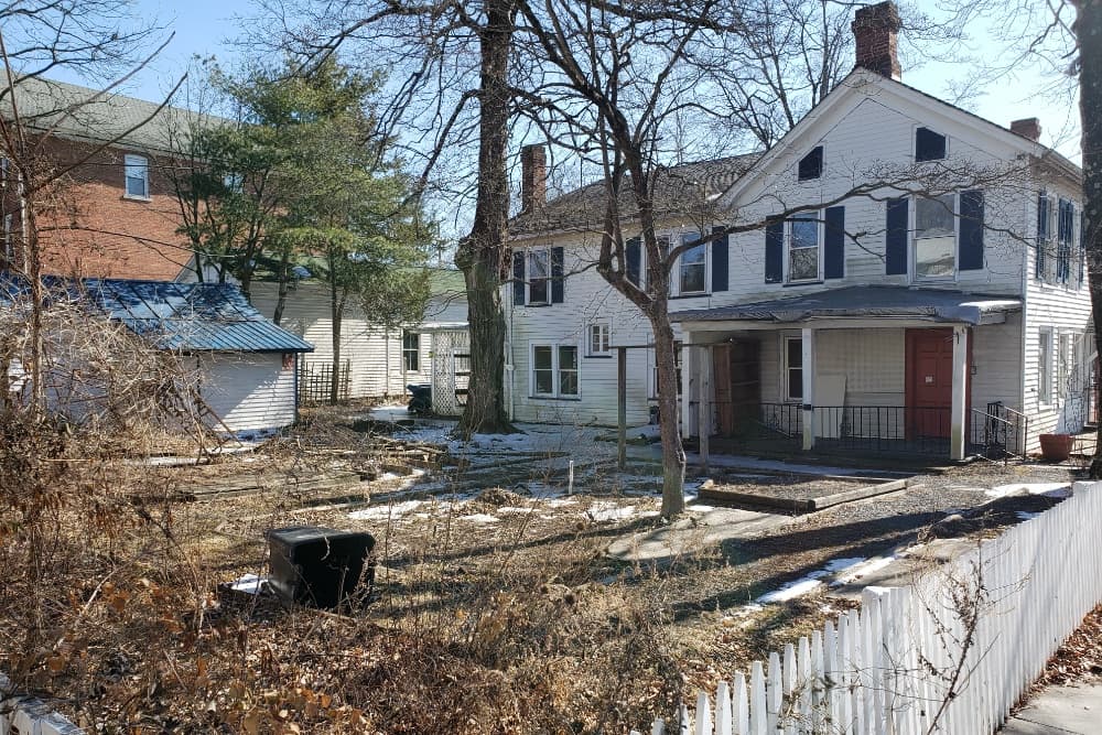 An old two-story house with a white picket fence and an overgrown yard, partially covered in snow.