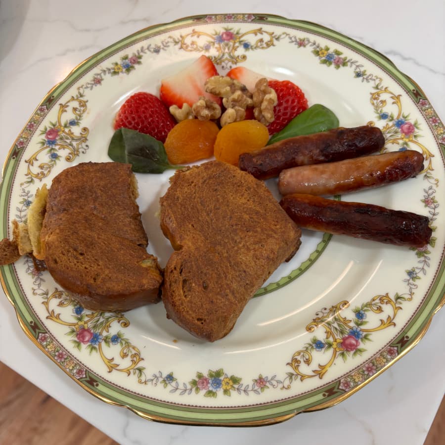 A plate of breakfast featuring two pieces of toast, sausages, and a side of strawberries, apricots, walnuts, and greens.