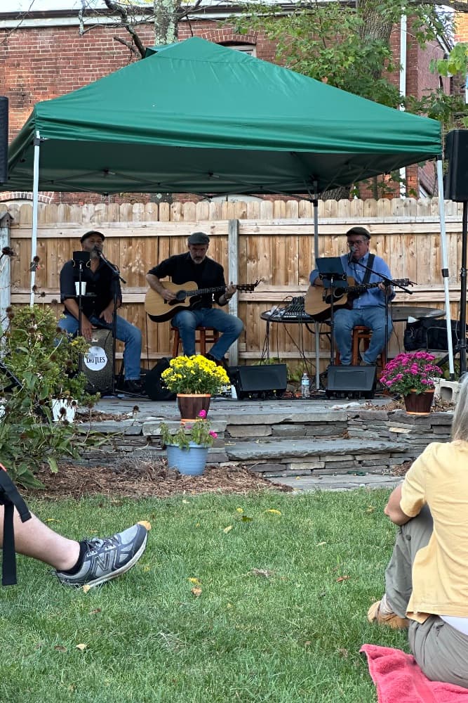 Three musicians perform under a green tent in a backyard garden.