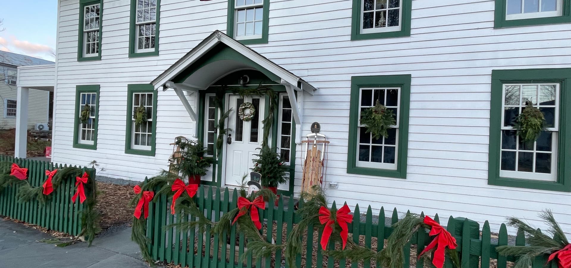 A festive white house decorated with green trim, wreaths, and red bows along the fence.