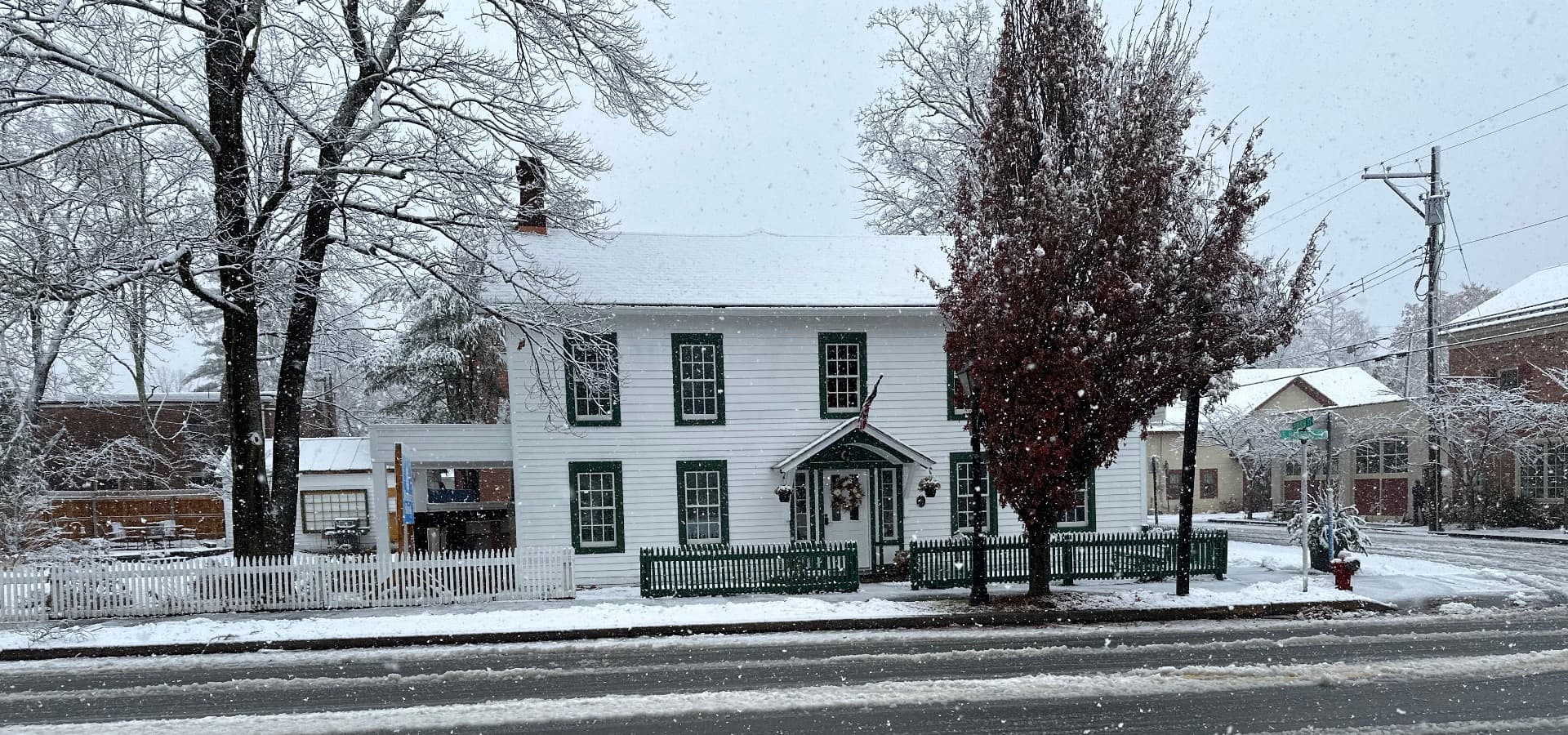 A historic white house with green shutters is surrounded by falling snow in a wintry landscape.