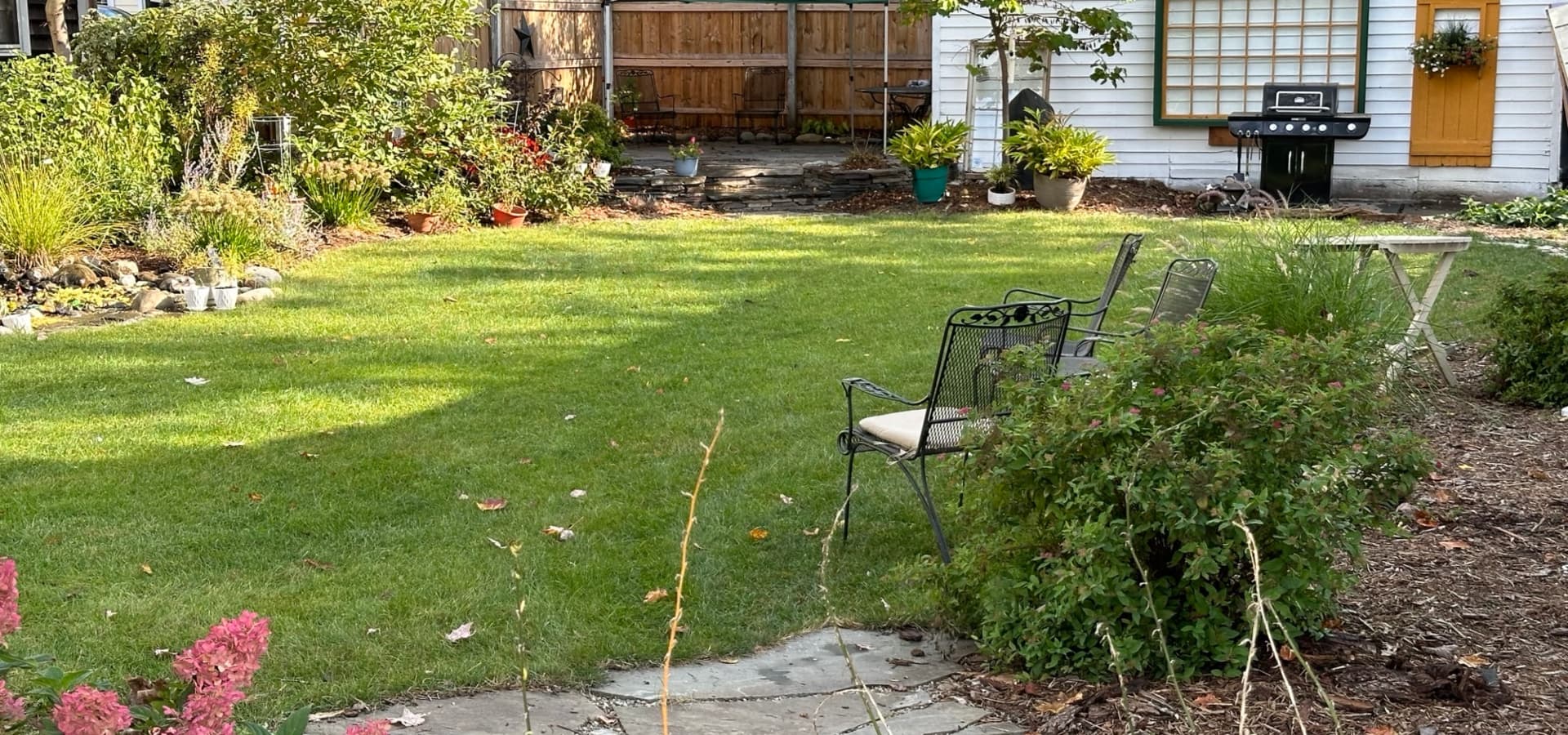 A lush backyard featuring a grassy area, flower beds, patio chairs, and a barbecue grill.