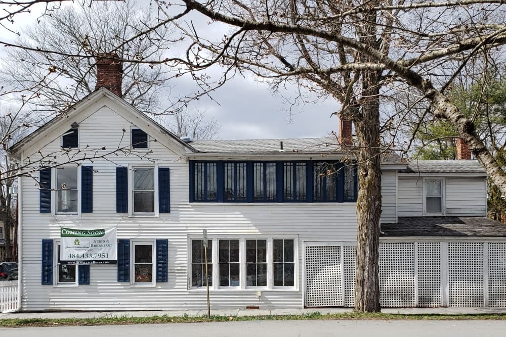 A white house with blue shutters and a "Coming Soon" sign for a bed and breakfast, surrounded by bare trees.