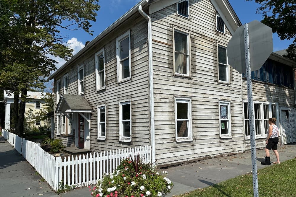 A weathered two-story wooden house with a porch, surrounded by a white picket fence and a woman walking nearby.