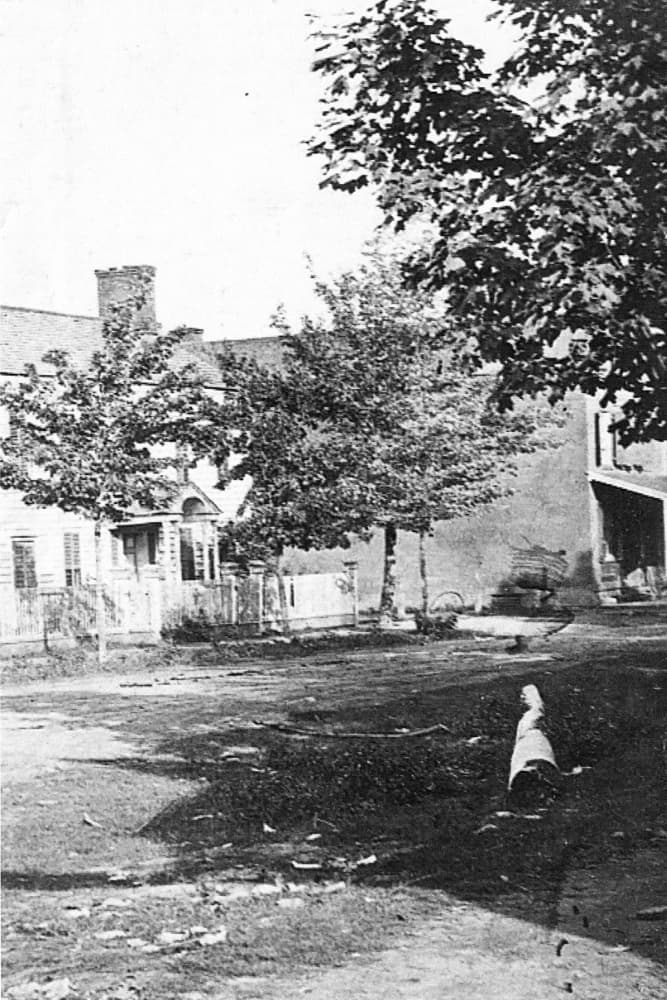 A historic black-and-white photograph of a yard with trees and a house in the background.